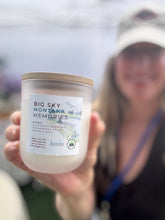 Load image into Gallery viewer, A hand holds a jar candle labeled 'BIG SKY MONTANA MEMORIES' in front of a blurred person wearing a white cap and dark top at what appears to be an outdoor market or event.
