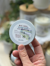 Load image into Gallery viewer, A hand holding a round tin of Big Sky Montana Memories, a travel candle, with a blurred background of similar jars and plants.