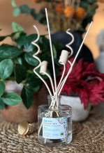 Load image into Gallery viewer, Diffuser in the Crisp Country Coeur d'Alene scent with decorative wood reeds on a table with foliage and flowers in the background