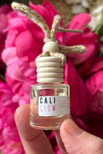 Load image into Gallery viewer, Car diffuser in a small apothecary bottle in the Cali Glow scent, held in a hand in front of a green leafy background.