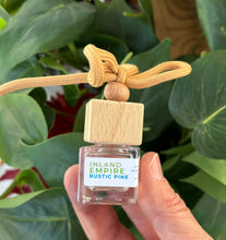 Load image into Gallery viewer, Mini glass jar with wooden lid as a car diffuser in the scent of "Inland Empire Rustic Pine" in front of greenery.