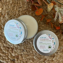 Load image into Gallery viewer, Two silver metal tin travel candles in the scent, "Crisp Country Coeur d'Alene," arranged open and closed on a woven surface with decorative fall leaves in the background.