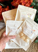 Load image into Gallery viewer, 4 Packages of Wax Melts in white packaging with window being held in front of orange fall flowers.