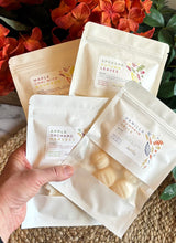 Load image into Gallery viewer, 4 Packages of Wax Melts in white packaging with window being held in front of orange fall flowers.