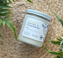 Load image into Gallery viewer, Glass candle jar labeled with scent 'Falling Leaves & Fisherman' on a woven surface with plants around.