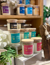 Load image into Gallery viewer, A shelf displaying various soy candles from Bungalow's Florida Collection with colorful labels, arranged in a storefront setting with a wooden background and flowers at the bottom.