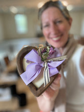 Load image into Gallery viewer, A person holding a heart-shaped wood dough bowl candle with a purple ribbon, eucalyptus embellishments, and a charm.