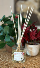 Load image into Gallery viewer, Glass reed diffuser jar in the Holiday Spice scent with wood reeds sitting on a table with green and red plants in background.