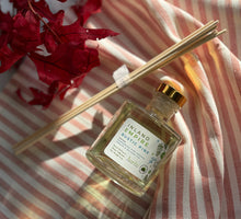 Load image into Gallery viewer, Glass reed diffuser jar in the Inland Empire Rustic Pine scent laying on a red stripped cloth with wood reeds next to it in a bundle.