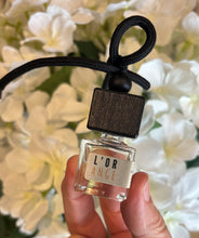 Load image into Gallery viewer, Bungalow car diffuser in the L'Orange scent held in front of white flowers.
