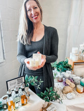 Load image into Gallery viewer, Sue holding massage serum candle with box at a booth event.