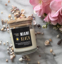 Load image into Gallery viewer, Candle in clear jar with wooden lid in the scent of 'Miami Beach' from Bungalow's Florida Collection sitting on a marble table surrounded by seashells and pink flowers.