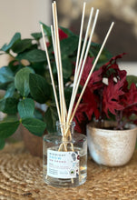 Load image into Gallery viewer, Glass reed diffuser jar in the Midnight Snow on Grand scent with wood reeds sitting on a table with green and red plants in background.