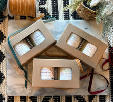 Load image into Gallery viewer, Three sets of two candles in brown window boxes with labels indicating different Fall scents, arranged on a table with a decorative bow and a woven basket in the background.