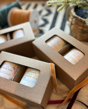 Load image into Gallery viewer, A set of two candles in brown window boxes with labels indicating different Fall scents, arranged on a table with a decorative bow and a woven basket in the background.