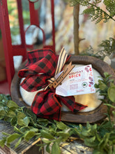 Load image into Gallery viewer, A hand-carved oval wood dough bowl candle with a checkered red and black ribbon and a 'Holiday Spice' labeled tag, adorned with green leaves in the background.