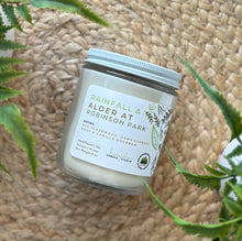 Load image into Gallery viewer, Glass candle jar labeled with scent 'Rainfall & Alder at Robinson Park' on a woven surface with plants around.