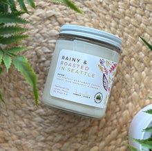 Load image into Gallery viewer, Glass candle jar labeled with scent 'Rainy & Roasted in Seattle' on a woven surface with plants around.
