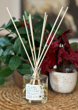 Load image into Gallery viewer, Glass reed diffuser jar in the Inland Empire Rustic Pine scent with wood reeds sitting on a table with green and red plants in background.