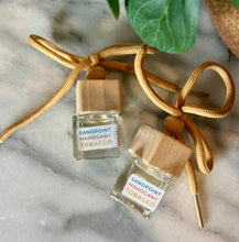 Load image into Gallery viewer, Two small car diffuser bottles with wooden caps in the scent of Sandpoint Mahogany Tobacco, lying on a marble surface.