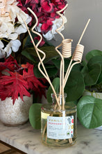 Load image into Gallery viewer, A reed diffuser bottle with a clear glass jar and curly, natural wood reeds. The white scent label reads, 'Vanilla Peppermint.' The bottle is set on a marble surface with red and white flowers and green leaves in the background.