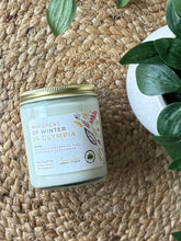 Load image into Gallery viewer, Glass candle jar labeled with scent 'Whispers of Winter in Olympia' on a woven surface with plants around.