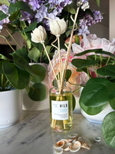 Load image into Gallery viewer, A reed diffuser bottle with white tulip-shaped reeds and a label showing the candle scent, Wild Surf, sits on a marble surface surrounded by potted plants and shells.