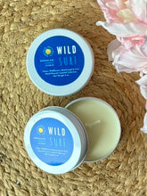 Load image into Gallery viewer, Two travel candle tins with a blue label in the Wild Surf scent on a woven surface with flowers.
