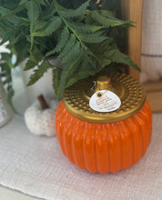 Load image into Gallery viewer, Orange pumpkin shaped candle with gold lid sitting on a textured mat with green foliage around it.