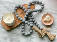 Load image into Gallery viewer, A Mango Wood Contentment Bead string, hand-painted in Gray, with jute tassels, draped next to a candle as home decor.