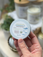 Load image into Gallery viewer, A hand holding a round white tin of 'Mountain Mornings in Whitefish' scented soy wax travel candle. The background is a soft-focus, warm-toned setting with other candles and decorative elements.

