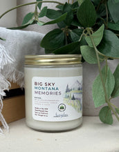 Load image into Gallery viewer, A glass jar candle with a gold metal lid and a white label reading 'BIG SKY MONTANA MEMORIES' styled on a tabletop with green trailing leaves and a white textured fabric in the background.
