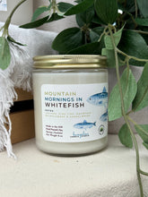 Load image into Gallery viewer, A jar candle labeled 'Mountain Mornings in Whitefish' sits on a light fabric surface, surrounded by green leaves and a white textured blanket. The candle has a white soy wax fill, a gold metal lid, and a white label with blue fish illustrations.
