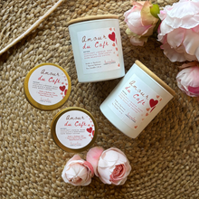 Load image into Gallery viewer, Two larger Amour du Cafe candles in white jars and two smaller candles in gold tin with decorative labels on a textured surface with pink flowers.
