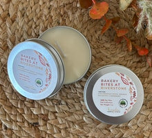 Load image into Gallery viewer, Two silver metal tin travel candles in the scent, &quot;Bakery Bites at Riverstone,&quot; arranged open and closed on a woven surface with decorative fall leaves in the background.
