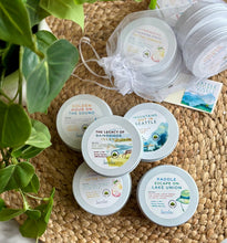 Load image into Gallery viewer, Collection of Seattle-inspired soy travel candles in a mesh bag as a bundle on a woven mat with a plant in the background.
