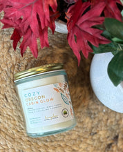 Load image into Gallery viewer, Candle labeled 'Cozy Oregon Cabin Glow' with red flowers in the background on a woven mat
