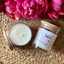 Load image into Gallery viewer, Two candles in glass jars with wooden lids sit on a woven surface with pink flowers-they're labeled with the scent of Cali Glow.
