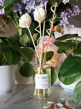 Load image into Gallery viewer, A reed diffuser bottle with white tulip-shaped reeds and a label showing the candle scent, CALI GLOW, sits on a marble surface surrounded by potted plants and shells.

