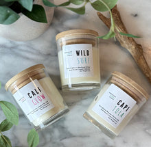 Load image into Gallery viewer, Three candles from the Beachy Coalstal Collection in clear glass jars and wooden lids on a marble surface with greenery
