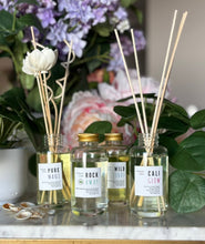 Load image into Gallery viewer, Four glass reed diffusers with natural wood reeds and white labels showing candle scents of Pure Maui, Rockaway, Wild Surf, and Cali Glow, styled with flowers and green leaves on a marble surface.
