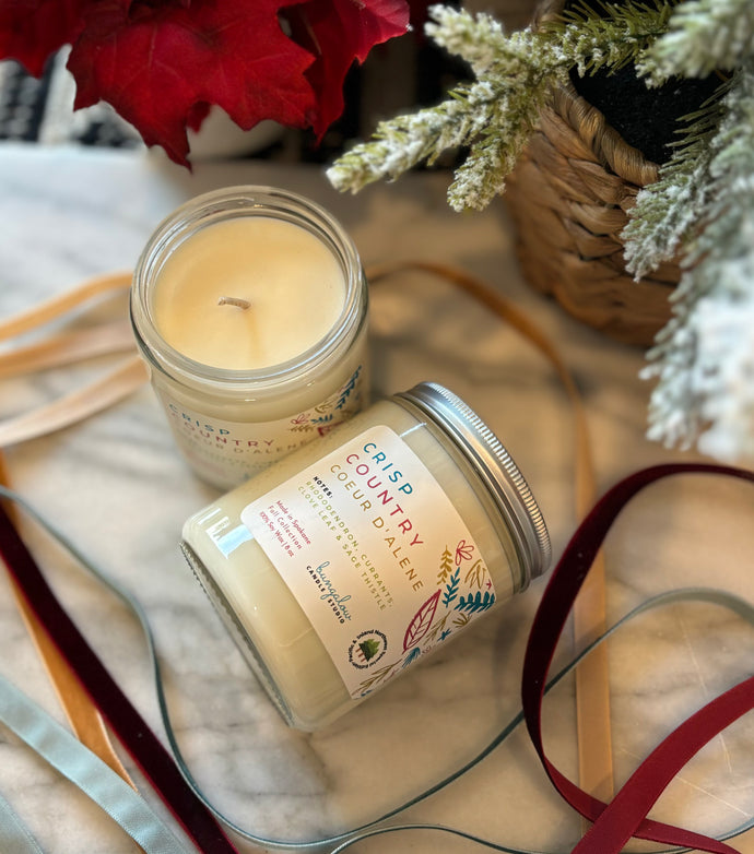 Two 8oz soy candles in glass jars with silver metal lids, labeled 'Crisp Country Coeur d'Alene'  placed on a marble surface with decorative red leaves and green branches in the background.