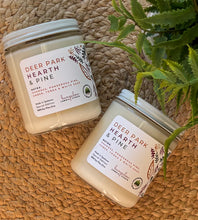 Load image into Gallery viewer, Two candles labeled &#39;Deer Park Hearth &amp; Pine&#39; on a woven surface with a plant.
