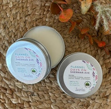 Load image into Gallery viewer, Two silver metal tin travel candles in the scent, &quot;Flannel Days Sherman Ave&quot; arranged open and closed on a woven surface with decorative fall leaves in the background.
