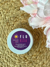 Load image into Gallery viewer, White travel candle tin with a purple label in the Florida scent on a woven surface with flowers.
