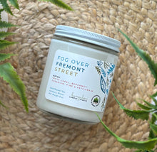 Load image into Gallery viewer, Glass candle jar labeled with scent &#39;Fog Over Fremont Street&#39; on a woven surface with plants around.
