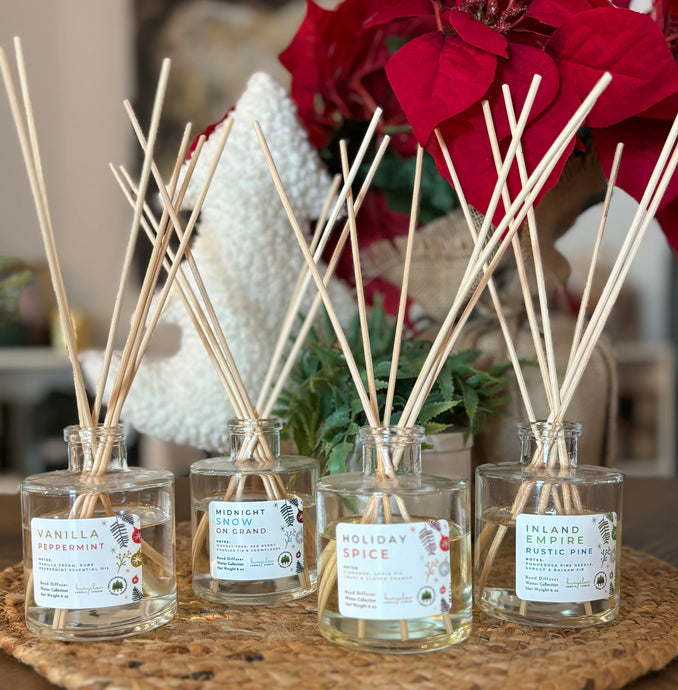 A group of clear glass reed diffusers with natural unbleached wood reeds, displaying various holiday scents such as vanilla peppermint, orange, and cinnamon.