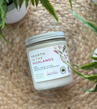 Load image into Gallery viewer, Candle jar labeled &#39;Hearth in the Highlands&#39; on a woven surface with plants around
