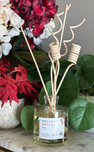 Load image into Gallery viewer, A clear glass reed diffuser bottle labeled 'Holiday Spice' with light natural curly reeds, styled on a marble surface with red and white flowers and green leaves in the background.
