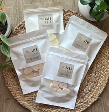 Load image into Gallery viewer, Four white sealed packages with clear windows showing heart shaped Wax Melts in the Lily Leaf, Lavender, Lemon, and Taos Sage scents, sitting on a textured mat with plants in background.
